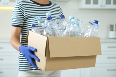 Woman holding cardboard box with used plastic bottles indoors, closeup. Recycling problem