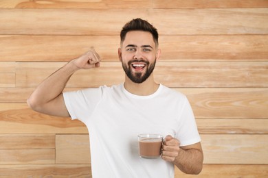 Young man with glass cup of chocolate milk showing his strength on wooden background