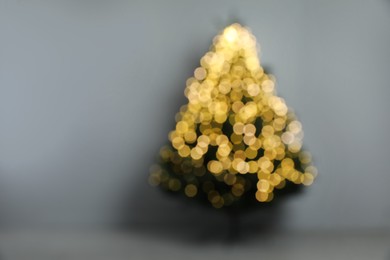 Blurred view of beautiful fir tree with Christmas lights  near grey wall in room, space for text