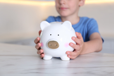 Little boy with piggy bank at marble table indoors, closeup