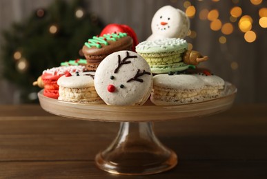 Stand with beautifully decorated Christmas macarons on wooden table against blurred festive lights