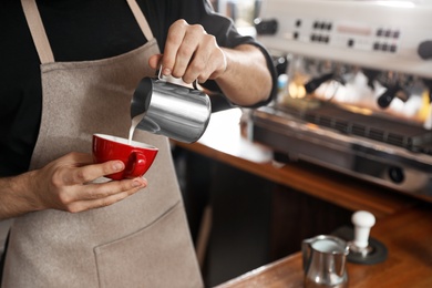 Photo of Barista pouring milk into cup of coffee in shop, closeup