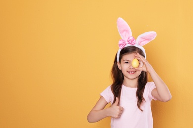 Little girl in bunny ears headband holding Easter egg near eye on color background, space for text
