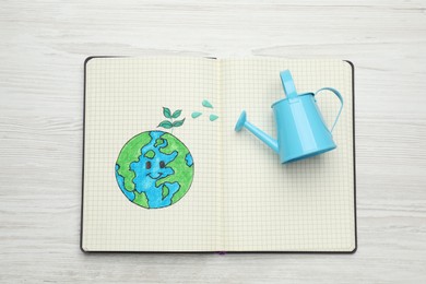 Happy Earth Day. Notebook with drawing of planet and watering can on white wooden table, top view