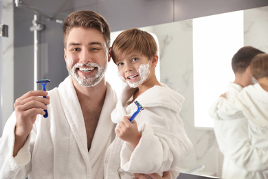 Dad and son with shaving foam on faces holding razors in bathroom