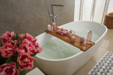 Wooden bath tray with candles and personal care products on tub indoors