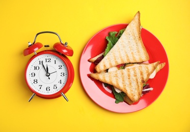 Photo of Tasty sandwiches and alarm clock on yellow background, flat lay. Nutrition regime