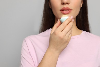 Young woman applying lip balm on grey background, closeup. Space for text
