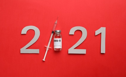 Paper numbers, syringe and vial with coronavirus vaccine forming 2021 on red background, flat lay