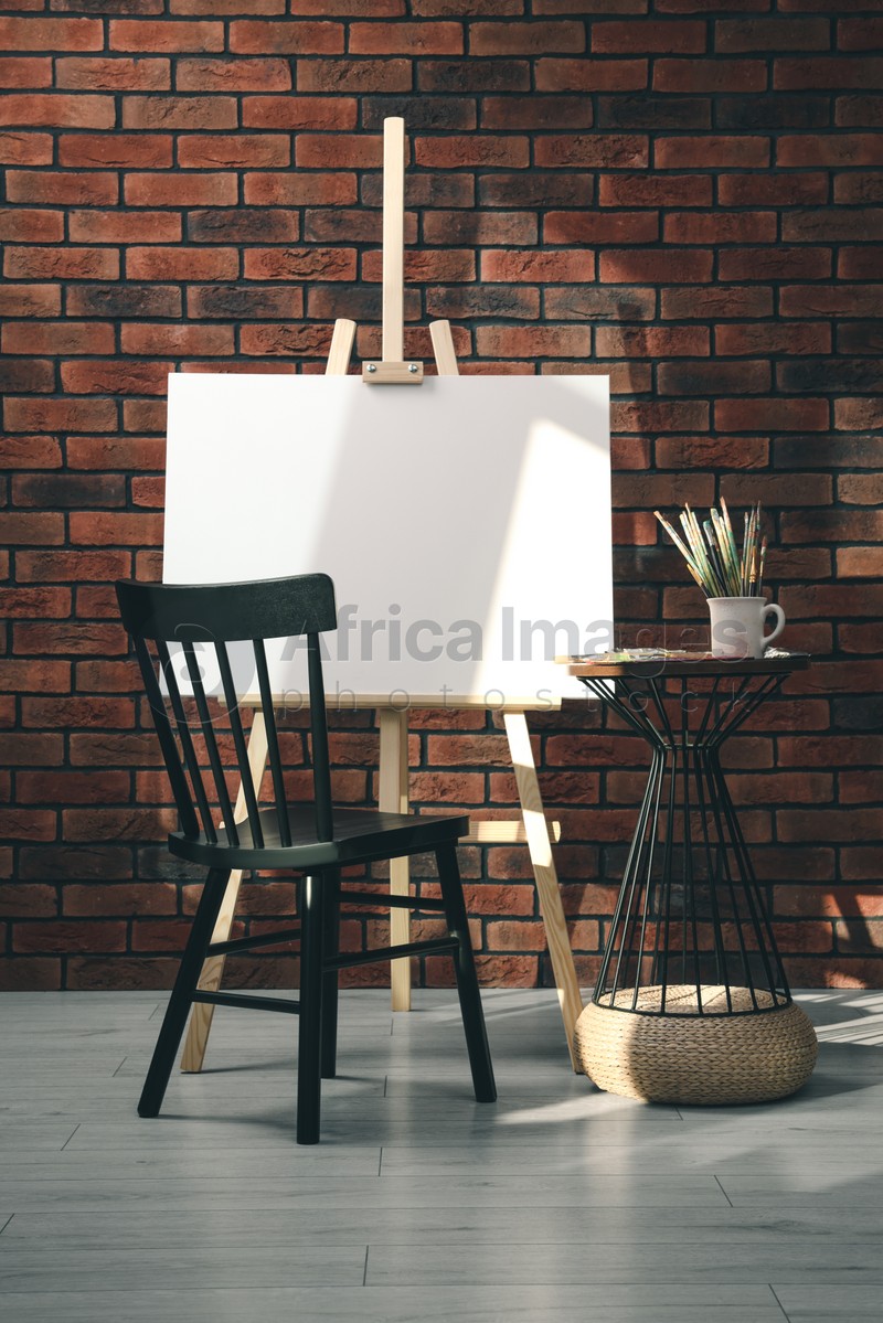 Photo of Wooden easel with blank canvas and different art supplies near brick wall in room