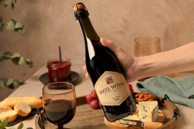 Photo of Woman holding bottle of red wine over table with different snacks, closeup