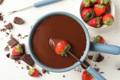 Dipping strawberry into fondue pot with chocolate on white table, top view