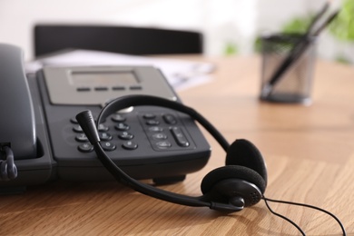 Desktop telephone and headset on wooden table in office, closeup. Hotline service