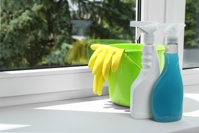 Green bucket with gloves and spray bottles of detergents on window sill indoors, space for text. Cleaning supplies