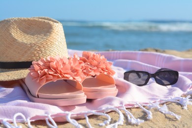 Photo of Blanket with stylish slippers, sunglasses and straw hat on sandy beach near sea, closeup