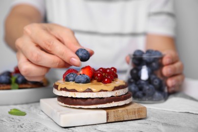 Woman decorating crunchy rice cakes with blueberry at white textured table, closeup