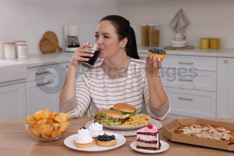Overweight woman drinking cola and holding cake in kitchen. Unhealthy food