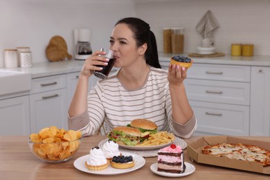 Photo of Overweight woman drinking cola and holding cake in kitchen. Unhealthy food