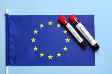 Test tubes with blood samples and European Union flag on light blue background, flat lay. Coronavirus outbreak