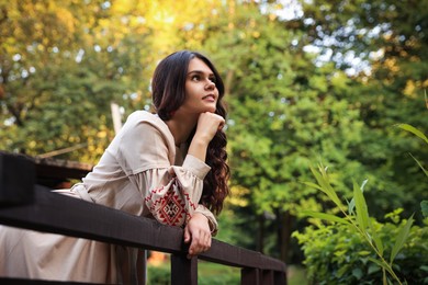 Beautiful woman wearing embroidered dress near wooden railing in countryside, space for text. Ukrainian national clothes