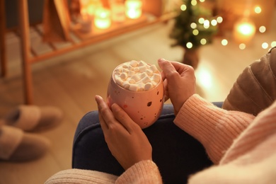 Woman holding cup of hot drink with marshmallows at home, closeup. Magic Christmas atmosphere