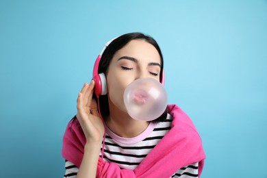 Fashionable young woman with headphones blowing bubblegum on light blue background