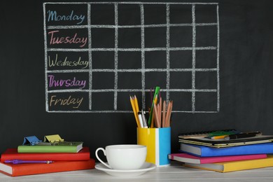 Cup of drink and different stationery on white wooden table near blackboard with chalked school timetable