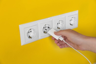 Woman plugging charger into power socket on yellow wall, closeup. Electrical supply