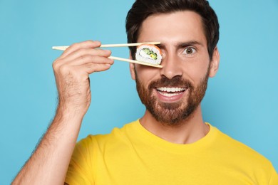 Happy man hiding his eye with tasty sushi roll on light blue background