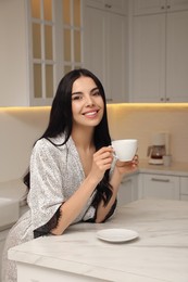 Pretty young woman wearing beautiful silk robe with cup of coffee in kitchen