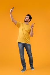 Photo of Smiling man taking selfie with smartphone and showing peace sign on yellow background