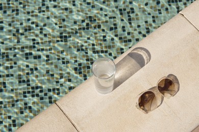 Stylish sunglasses and glass of water near outdoor swimming pool on sunny day, above view. Space for text