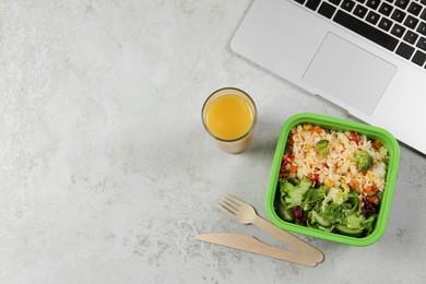 Photo of Container of tasty food, laptop, cutlery and glass of juice on light grey table, flat lay with space for text. Business lunch