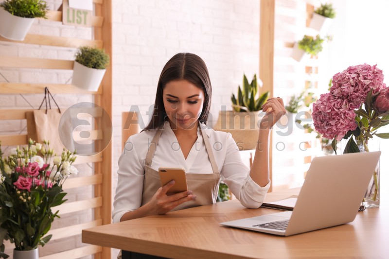 Photo of Florist with smartphone working on laptop in workshop
