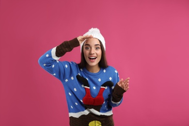 Surprised young woman in Christmas sweater and hat on pink background