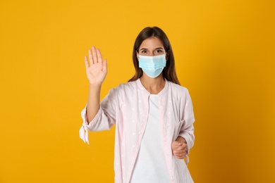 Woman in protective mask showing hello gesture on yellow background. Keeping social distance during coronavirus pandemic