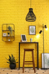 High wooden table with tablet and stool as stand up workplace near yellow brick wall. Stylish interior