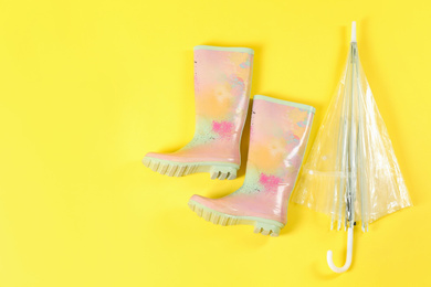 Transparent umbrella and colorful rubber boots on light yellow background, flat lay