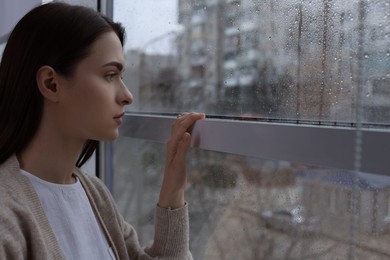 Melancholic young woman looking out of window on rainy day, space for text. Loneliness concept