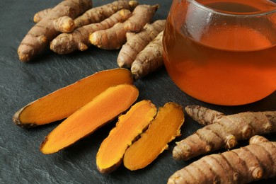 Photo of Glass cup of tasty tea, whole and cut turmeric roots on black textured table, closeup