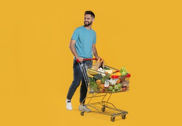 Happy man with shopping cart full of groceries on yellow background