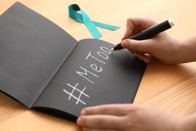 Woman writing hashtag MeToo in notebook near teal awareness ribbon at table, closeup. Stop sexual assault