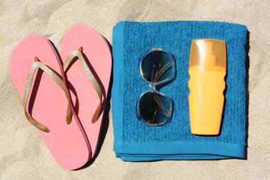 Folded soft blue beach towel with flip flops, sunglasses and bottle of sunscreen on sand, flat lay
