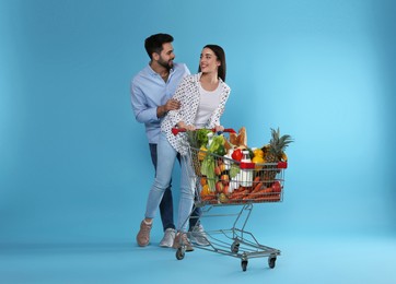 Young couple with shopping cart full of groceries on light blue background