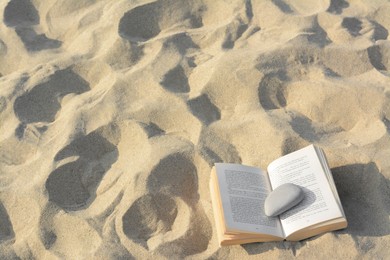 Open book with stone on sandy beach, above view. Space for text