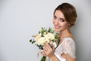 Young bride with elegant hairstyle holding wedding bouquet on light grey background. Space for text