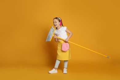 Photo of Cute little girl in headphones with broom singing on orange background