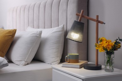 Stylish lamp, book, hot drink and flowers on bedside table indoors. Bedroom interior element