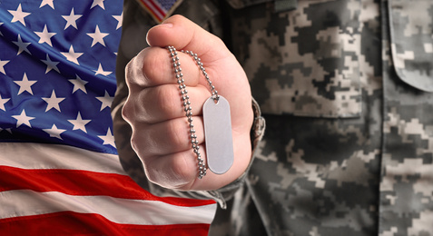 Male soldier holding military ID tag and American flag on background, closeup. Military service