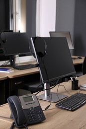 Modern computer with headset and stationary phone on wooden desk in office. Hotline service